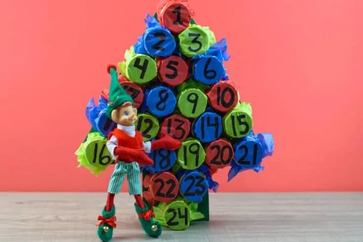 Christmas elf next to homemade advent calendar against pink backdrop