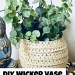 A wicker vase with faux greenery sits on a wooden surface next to a Buddha statue, a sun hat, and a beaded garland. Text reads "DIY Wicker Vase. Easy Dollar Tree DIY!.