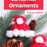 two handmade red Santa hat ornaments made from yarn laying on faux pine needles