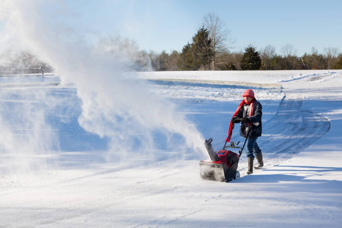 Senior lady using a snowblower on rural drive on windy day with a cloud or blizzard of snow blowing in the air