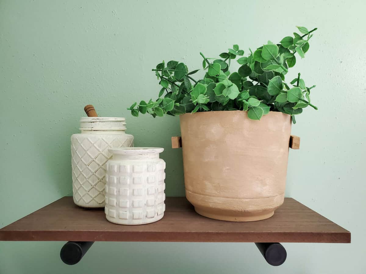A faux terra cotta pot with ivy plant on a brown shelf and 2 cream vases with a green wall behind.