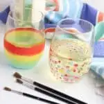 A colorful towel and wine bottle behind two painted wine glasses filled with white wine; with red, yellow, green, and blue stripes and polka dots with three clean paint brushes lying in front.