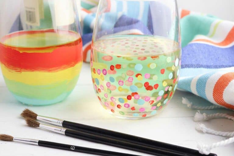 A colorful towel behind two painted wine glasses with red, yellow, green, and blue stripes and polka dots with three clean paint brushes lying in front.