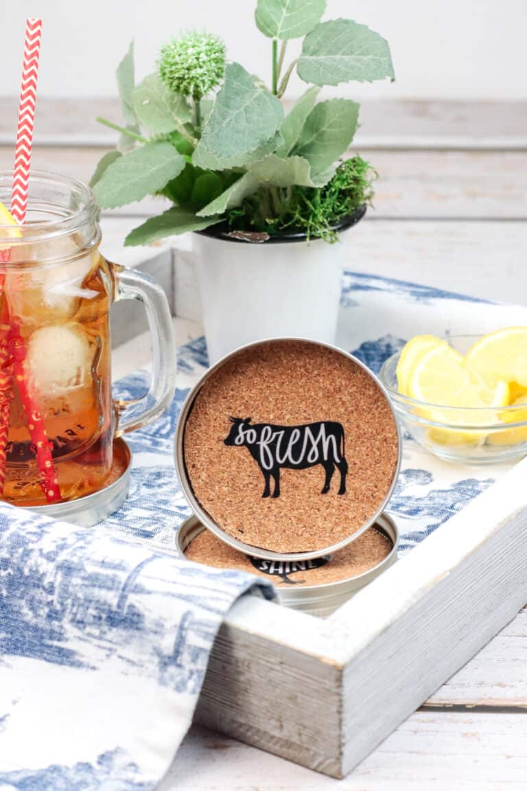 A serving try holds a small plant, a glass bowl of lemon wedges, a mason jar filled with iced sweet tea and a red striped straw sitting in a mason jar cupholder; a mason jar cupholder is propped up showing the farmhouse decal on the cork surface.