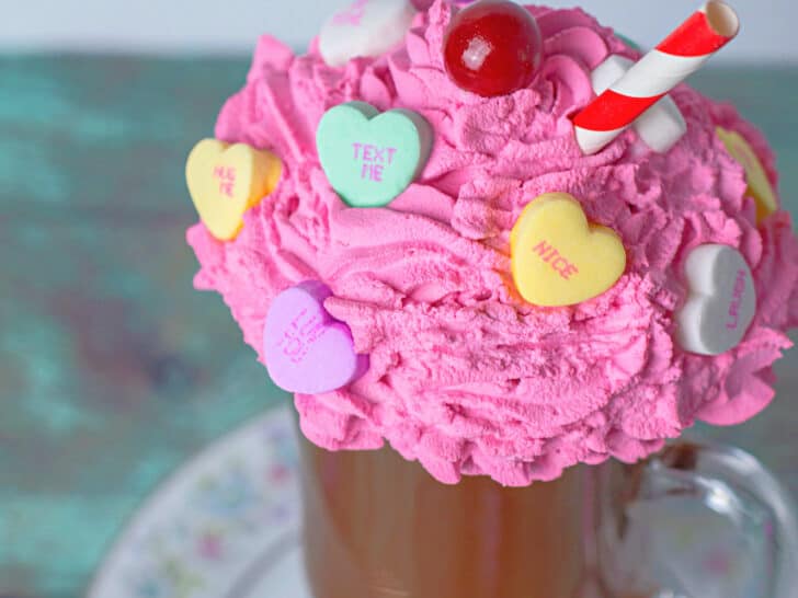 close up of a diy fake hot chocolate mug topped with pink whipped cream decorated with candy hearts and a red and white straw displayed on a china plate on top of a weathered wood table with candy hearts