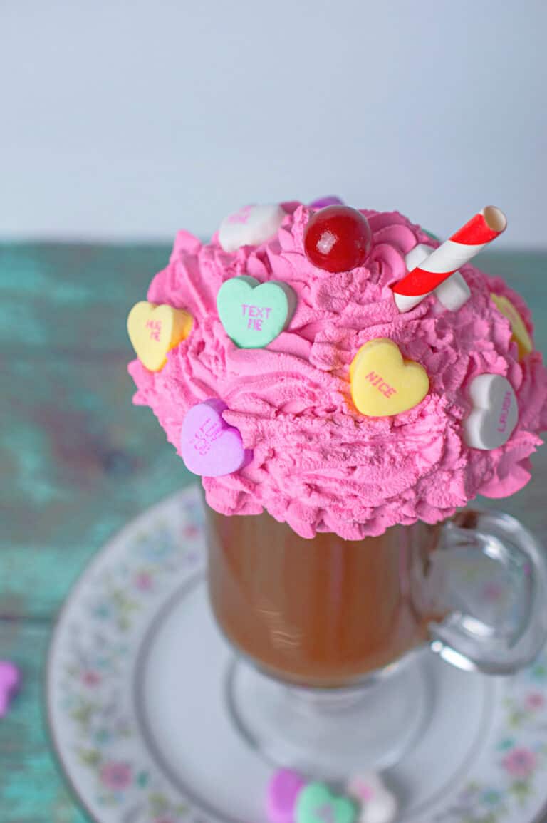 close up of a diy fake hot chocolate mug topped with pink whipped cream decorated with candy hearts and a red and white straw displayed on a china plate on top of a weathered wood table with candy hearts