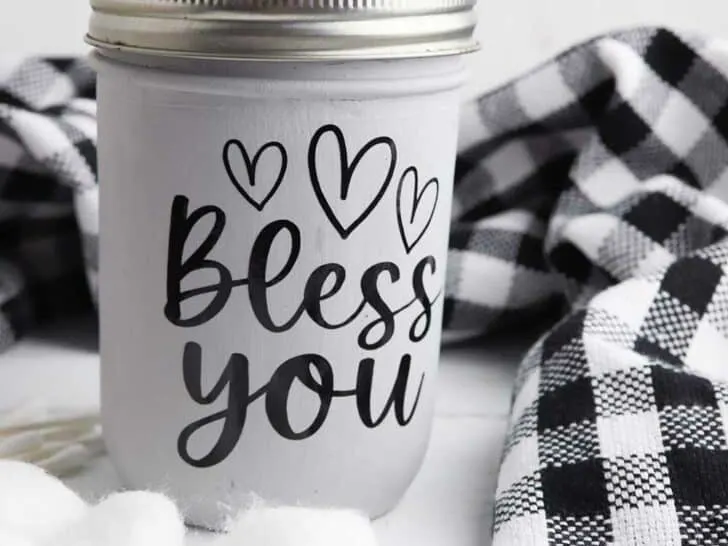Bless You mason jar tissue holder on a white counter top with black plaid towel and cotton ball laying nearby.