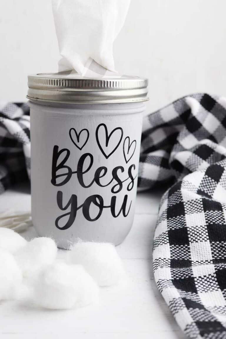 Bless You mason jar tissue holder on a white counter top with black plaid towel and cotton ball laying nearby.