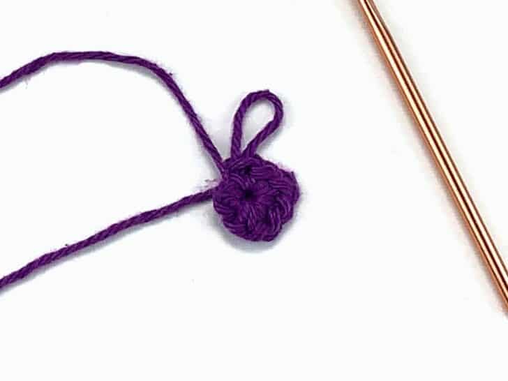 purple yarn and a gold crochet hook working a magic ring crochet stitch against a white background