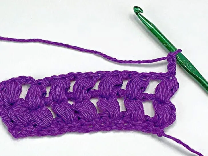 purple yarn and a green crochet hook working a puff stitch against a white background