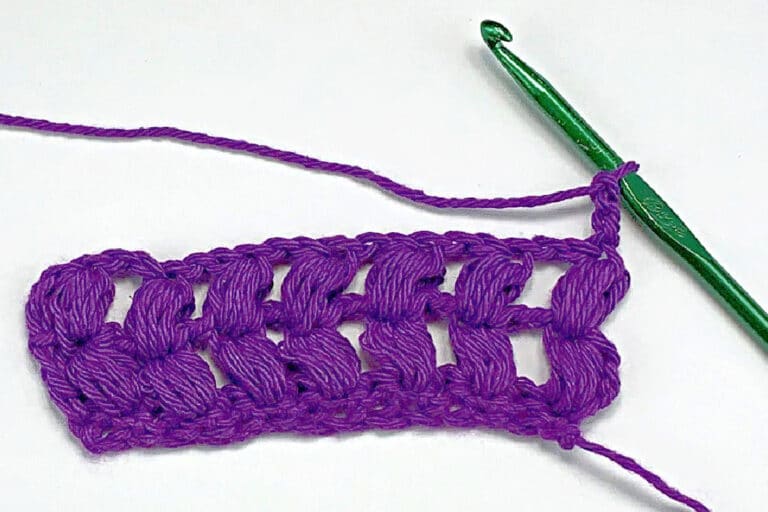 purple yarn and a green crochet hook working a puff stitch against a white background