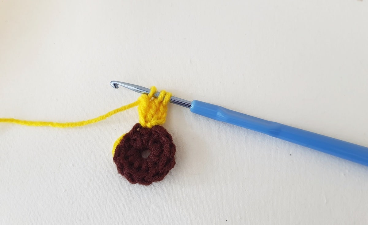 Crochet Sunflower Granny Square Step 10 A yellow and brown crocheted flower with a blue crochet hook.