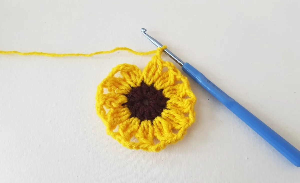 Crochet Sunflower Granny Square Step 16 A yellow crochet flower with a blue crochet hook.