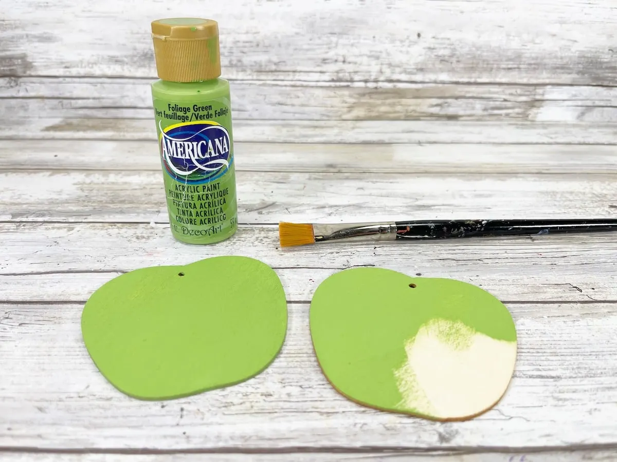 Candy Apple Craft Step 3 Two green apple ornaments and a paint brush on a wooden table.