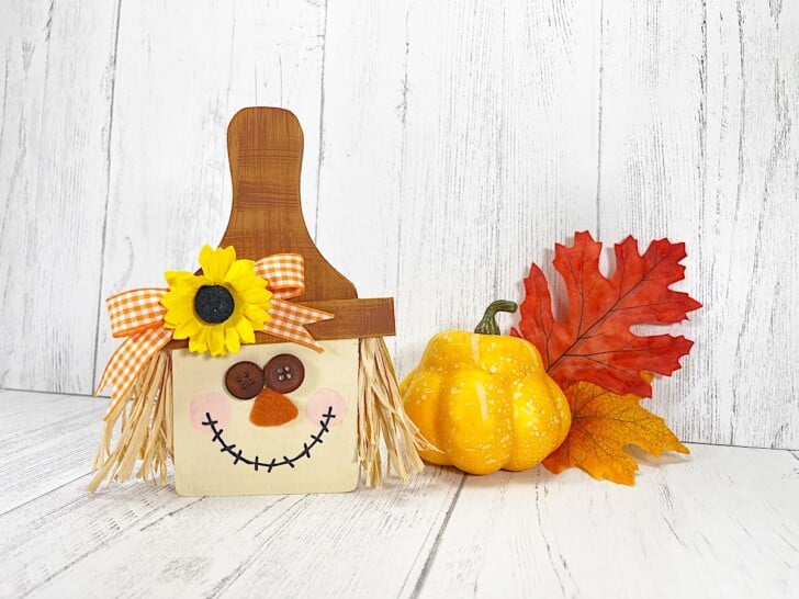 Cutting Board Scarecrow next to an orange pumpkin and red and yellow fall leaves.