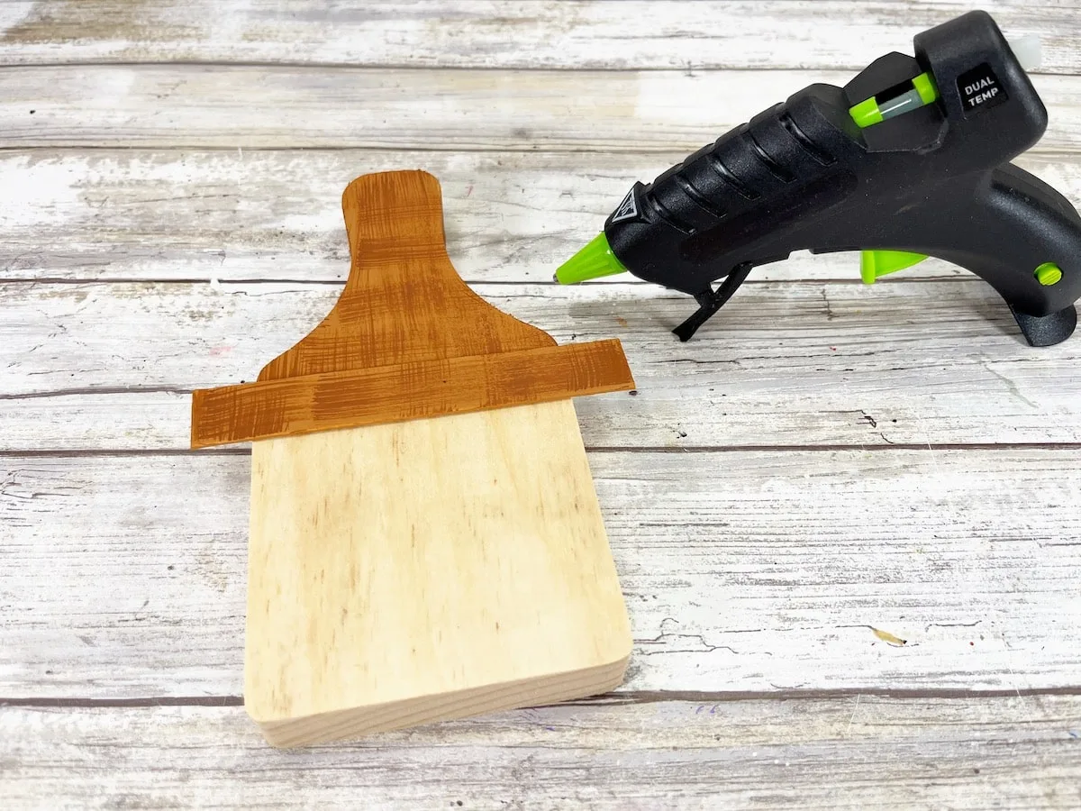 Cutting Board Scarecrow Step 5B with a glue gun next to a wooden cutting board.