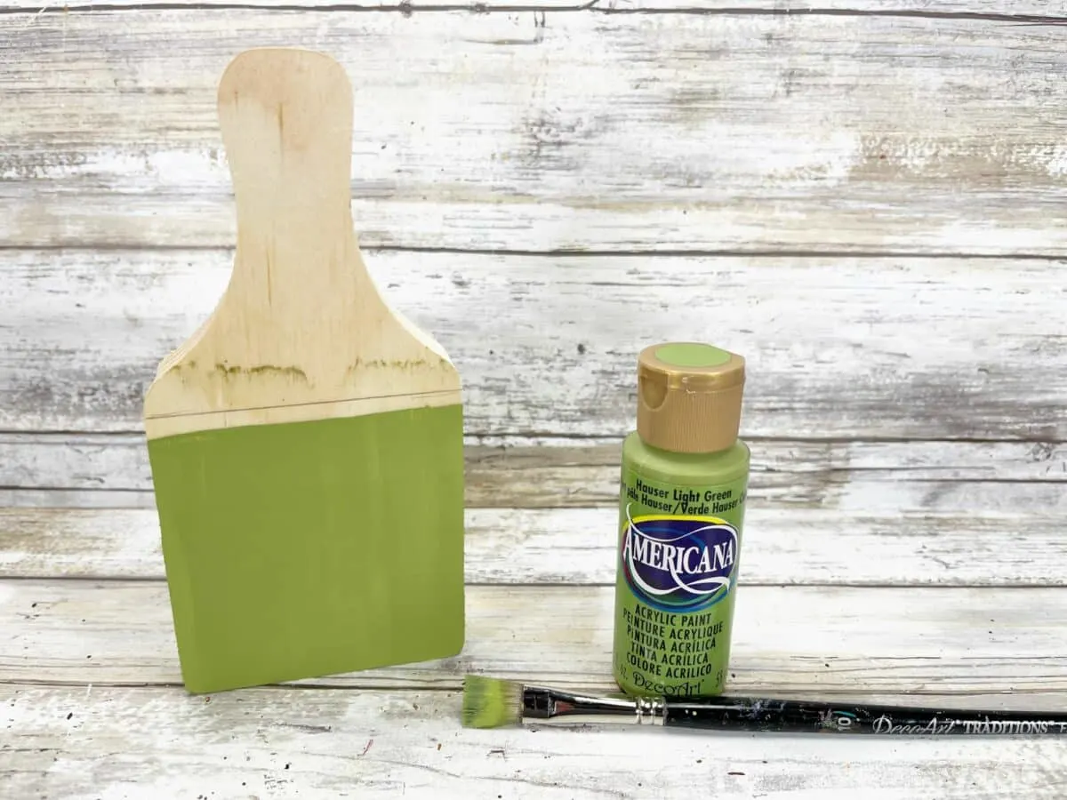 A green paint brush and a paint brush next to a wooden board.