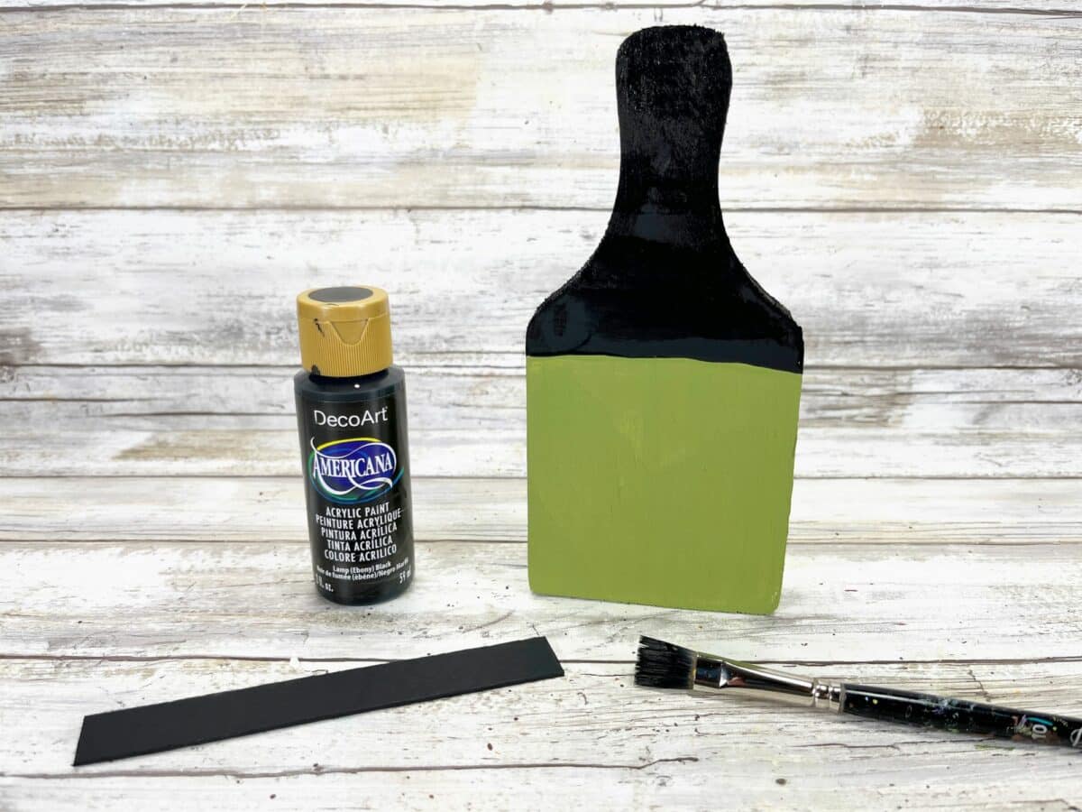 A green paint brush, black paint, and a black paint brush on a wooden table.