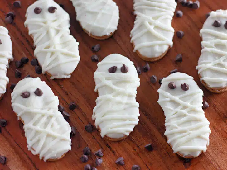 Mummy cookies decorated with white frosting and chocolate chip eyes are arranged on a wooden surface, surrounded by scattered chocolate chips.