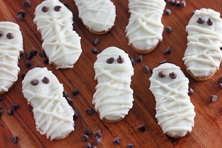 Mummy cookies decorated with white frosting and chocolate chip eyes are arranged on a wooden surface, surrounded by scattered chocolate chips.