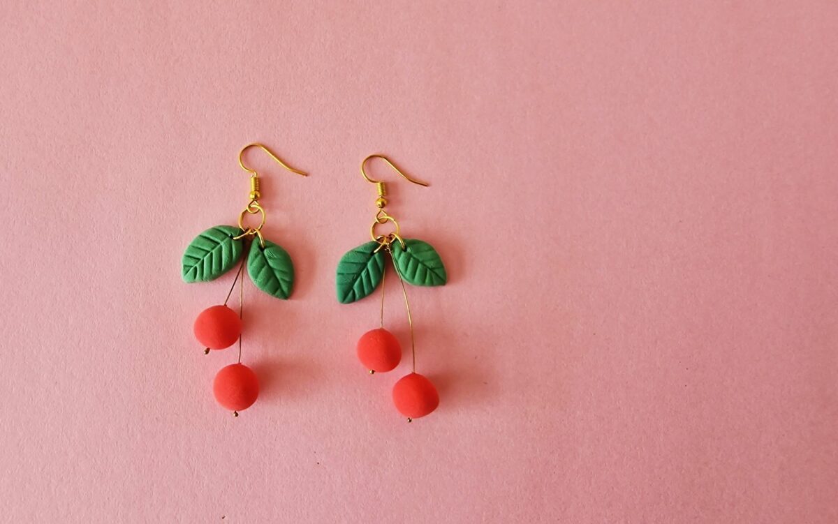 A pair of earrings with red cherries and leaves on a pink background.