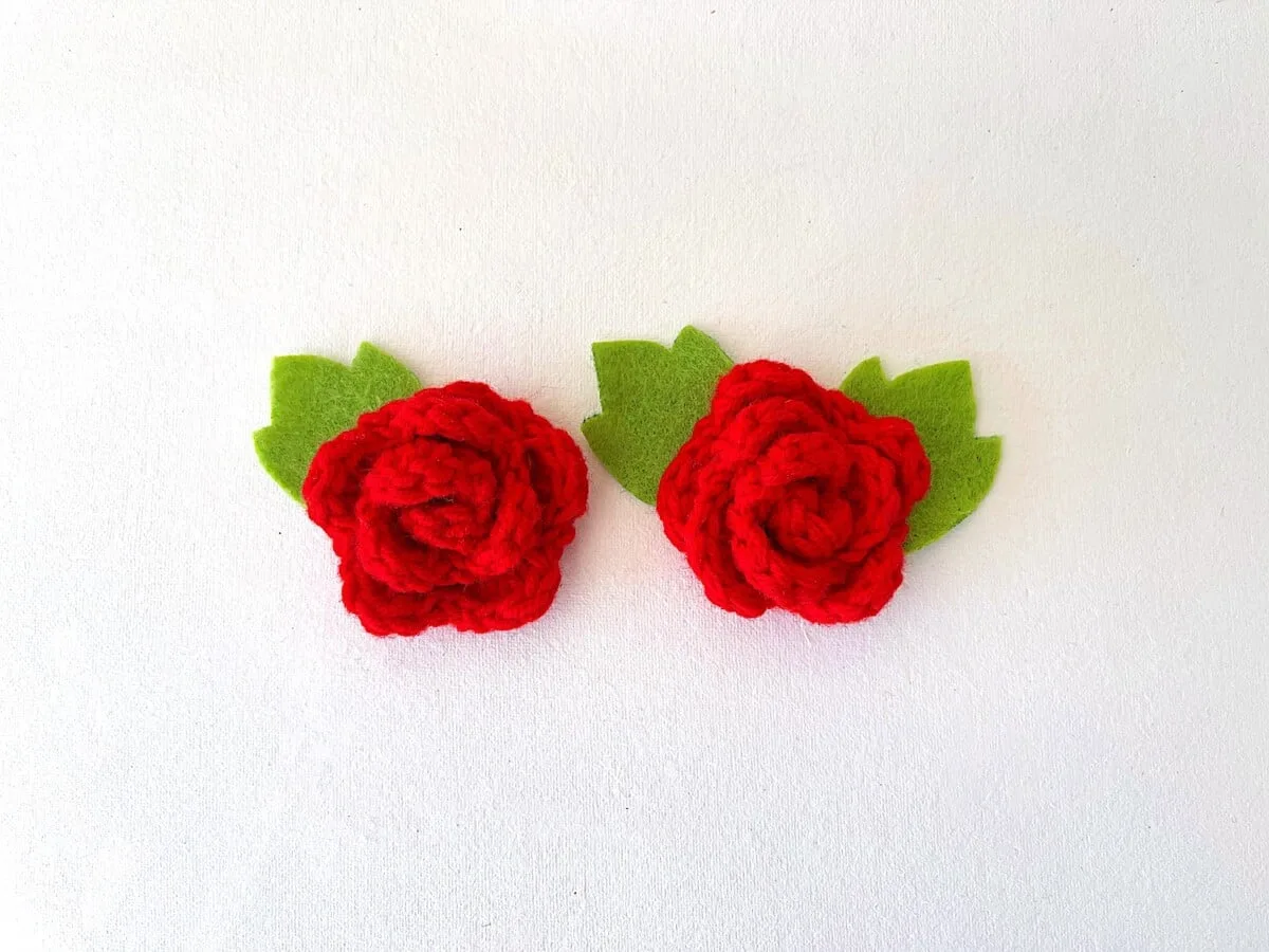 Two red crochet roses with green leaves on a white surface.
