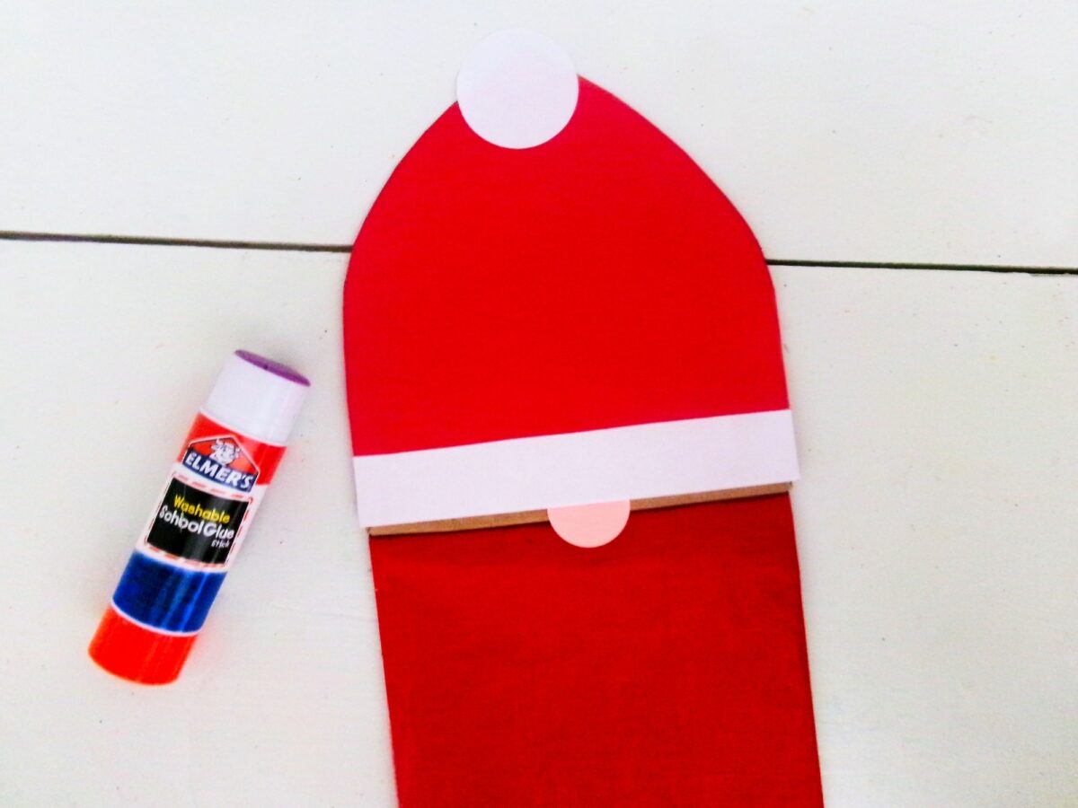 An adorable santa gnome is wearing a santa claus hat and holding a red tube of glue.
