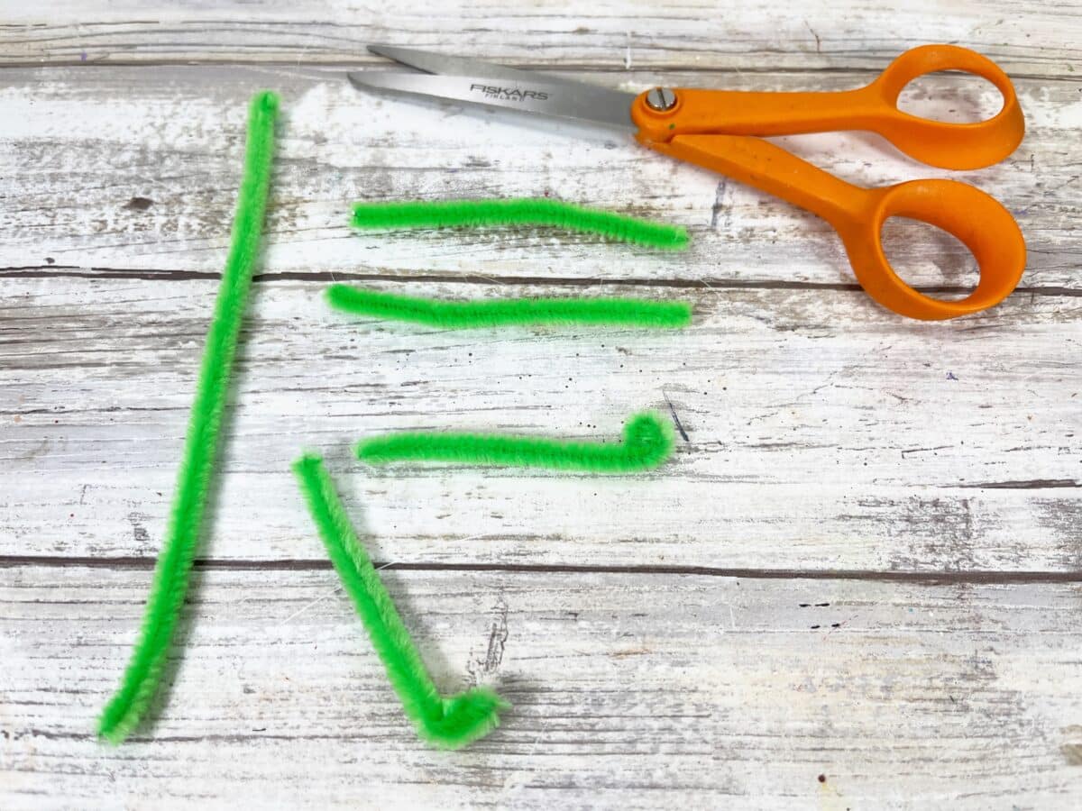 A pair of scissors on a wooden table next to a green craft stick.