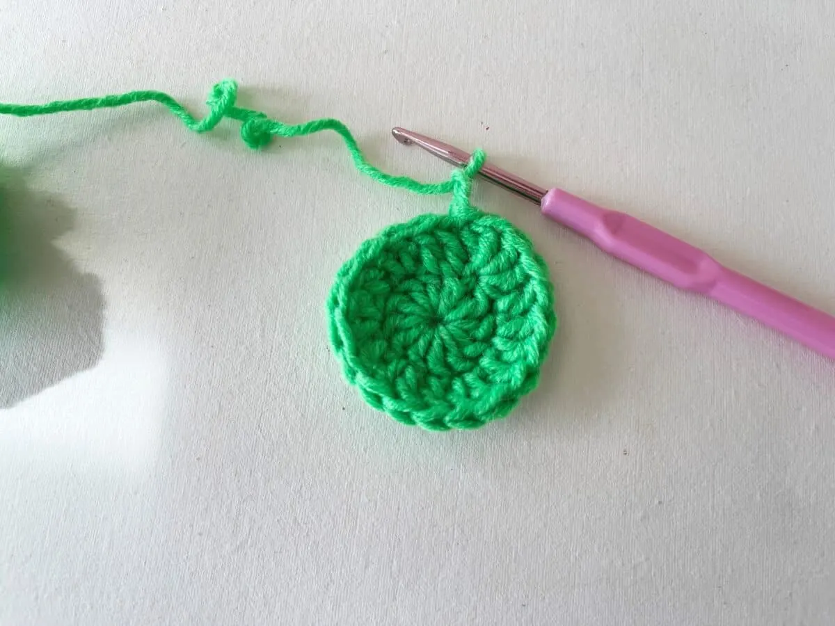 A green crocheted flower with a pink crochet hook.