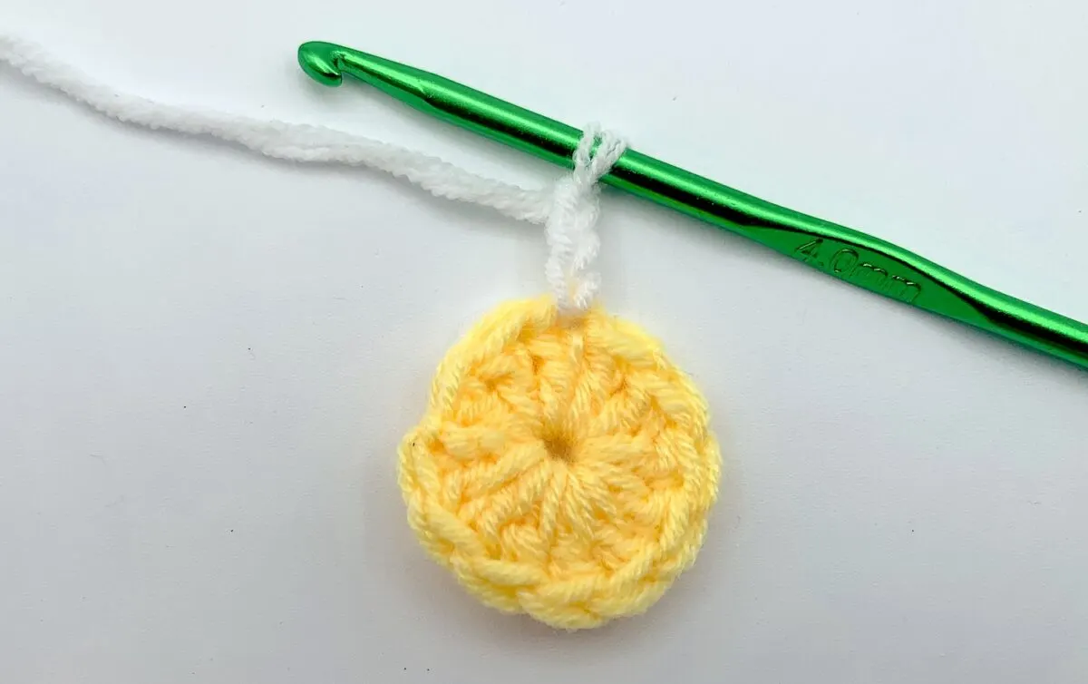 Round 2 - Step 1.2 A yellow crocheted flower with a green crochet hook.