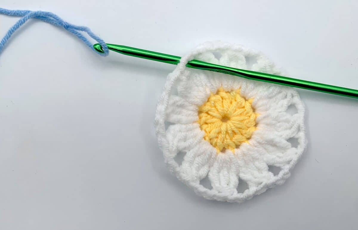 Round 3 - Step 1 A crocheted daisy with a green crochet hook.