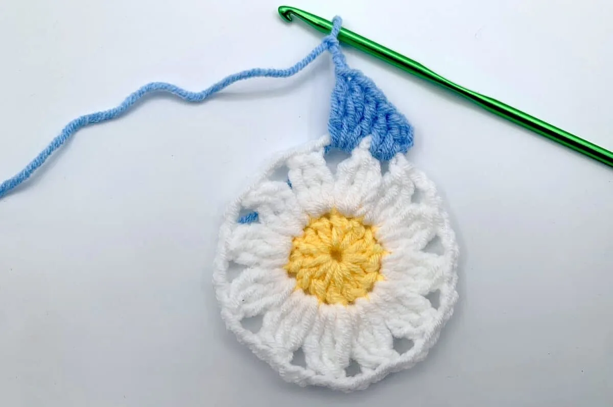 Round 3 - Step 5 A crocheted daisy with a blue and white crochet hook.