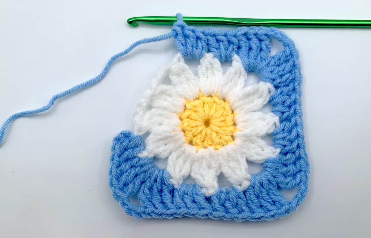 Round 3 - Step 11 A blue and white crochet square with a daisy on it.