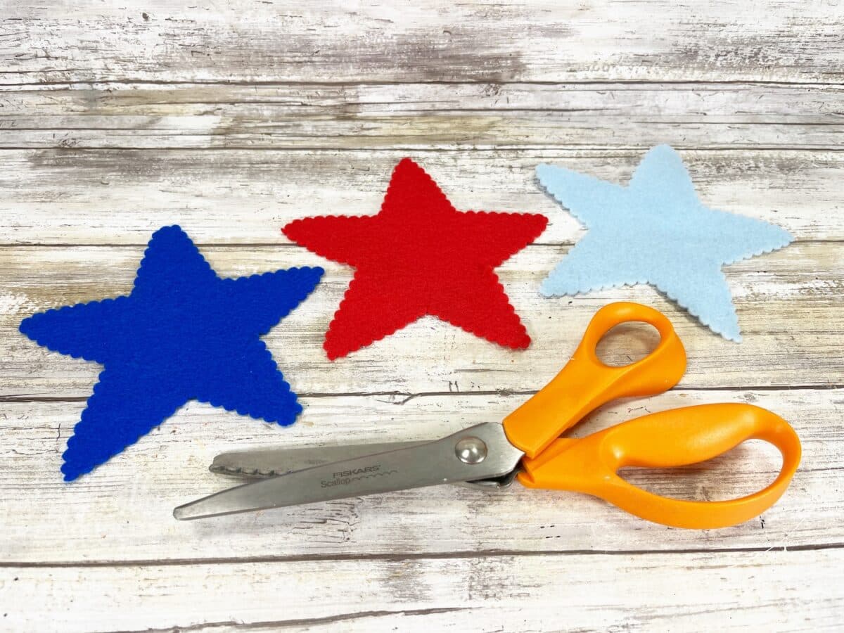 Felt Star People Step 2 Three felt stars in blue, red, and light blue alongside a pair of orange-handled scissors on a wooden surface.