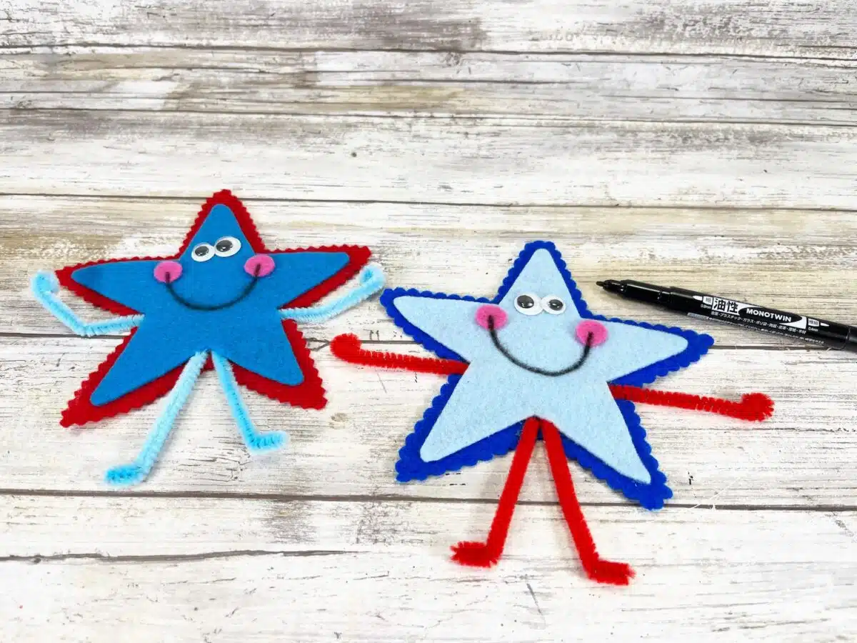 Felt Star People Step 8 Two homemade star-shaped crafts with googly eyes and pipe cleaner arms and legs on a wooden surface, with a marker lying nearby.