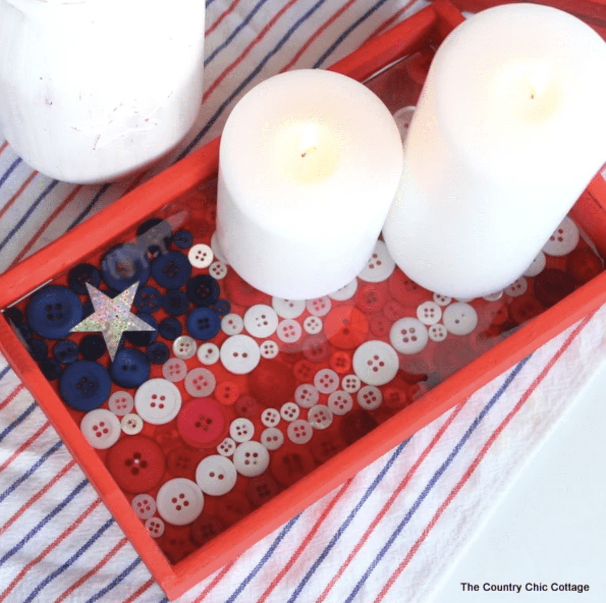 Two white candles placed in a red tray filled with an assortment of red, white, and blue buttons, perfect for Fourth of July crafts.