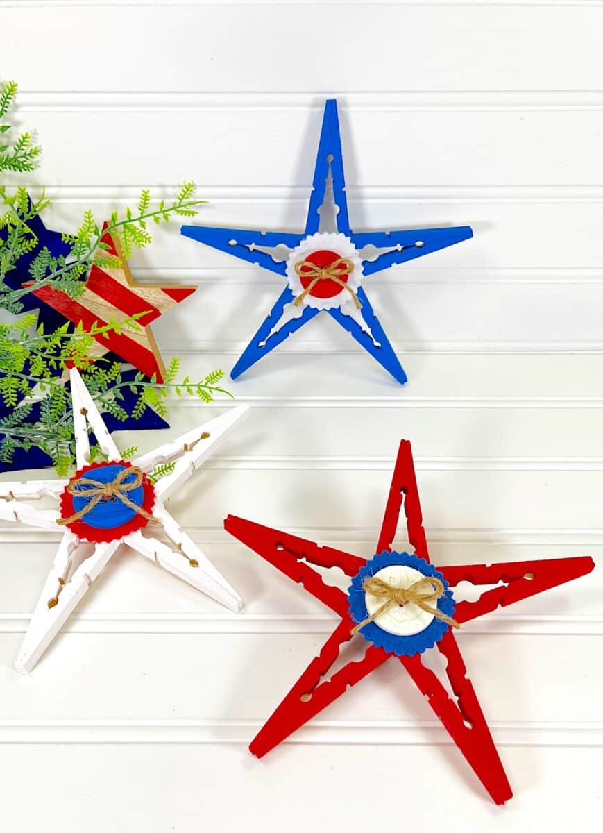Patriotic Clothespin Stars painted in red, white, and blue, displayed on a white background with greenery accents.