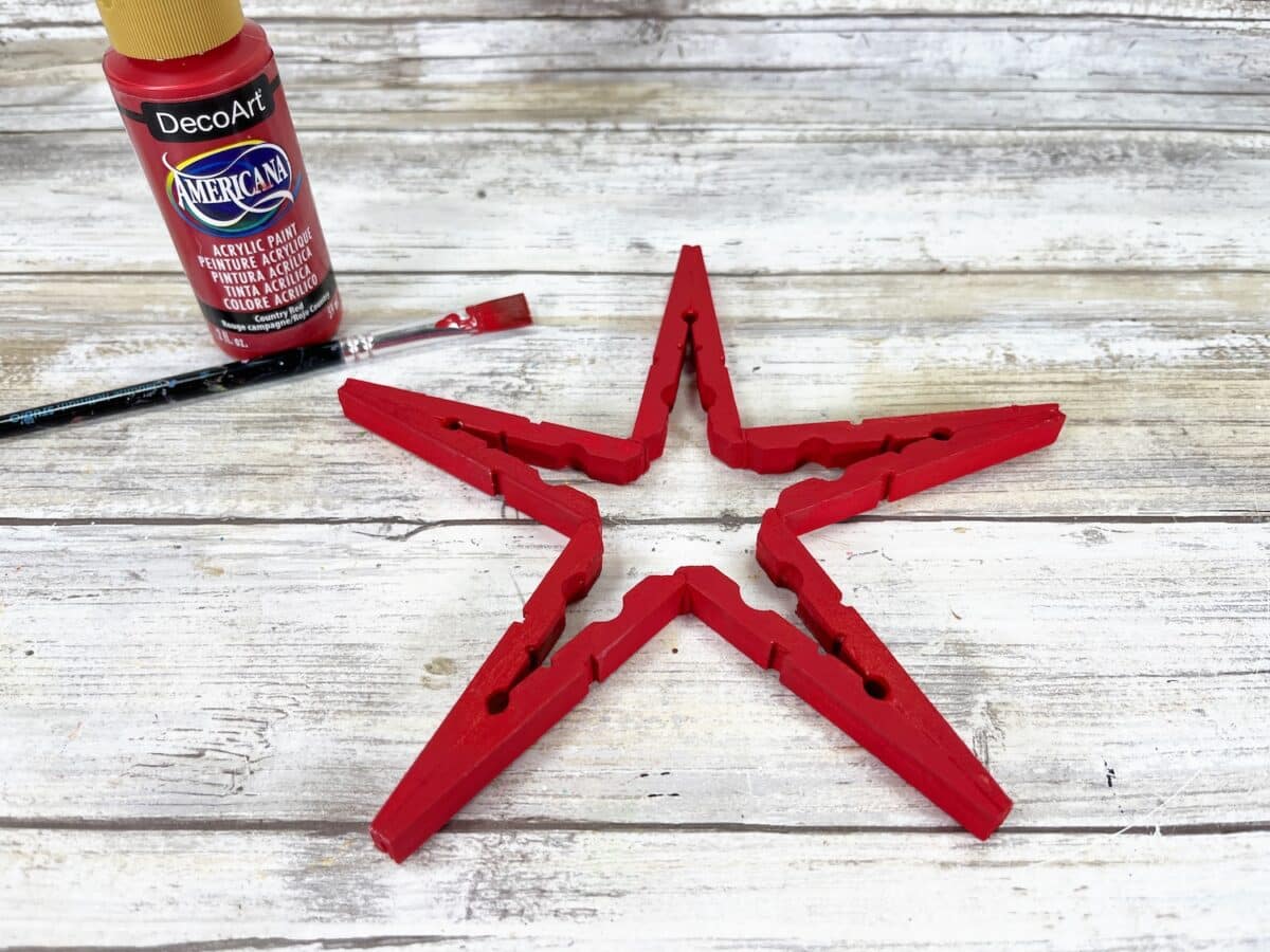 Patriotic Clothespin Stars Step 4 A red wooden star with a bottle of red acrylic paint and a paintbrush on a rustic white wooden background.