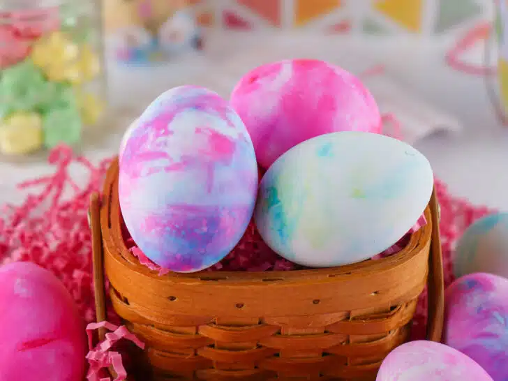 Colorful shaving cream eggs in a woven basket with festive decorations in the background.