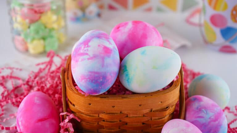 Colorful shaving cream eggs in a woven basket with festive decorations in the background.