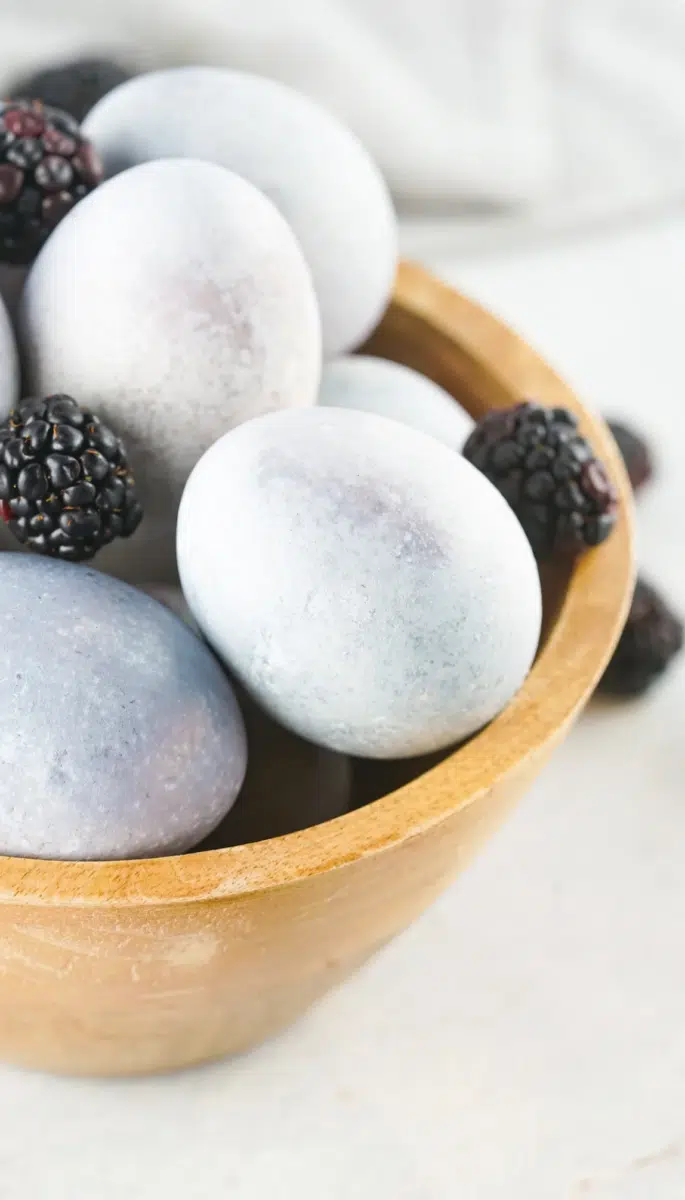 A wooden bowl containing frosted blue eggs dyed in cool ways and blackberries.