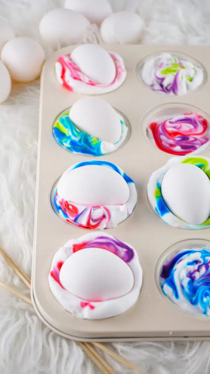 White eggs positioned in a muffin tin with several cups containing shaving cream eggs dyed with swirls of colorful paint.