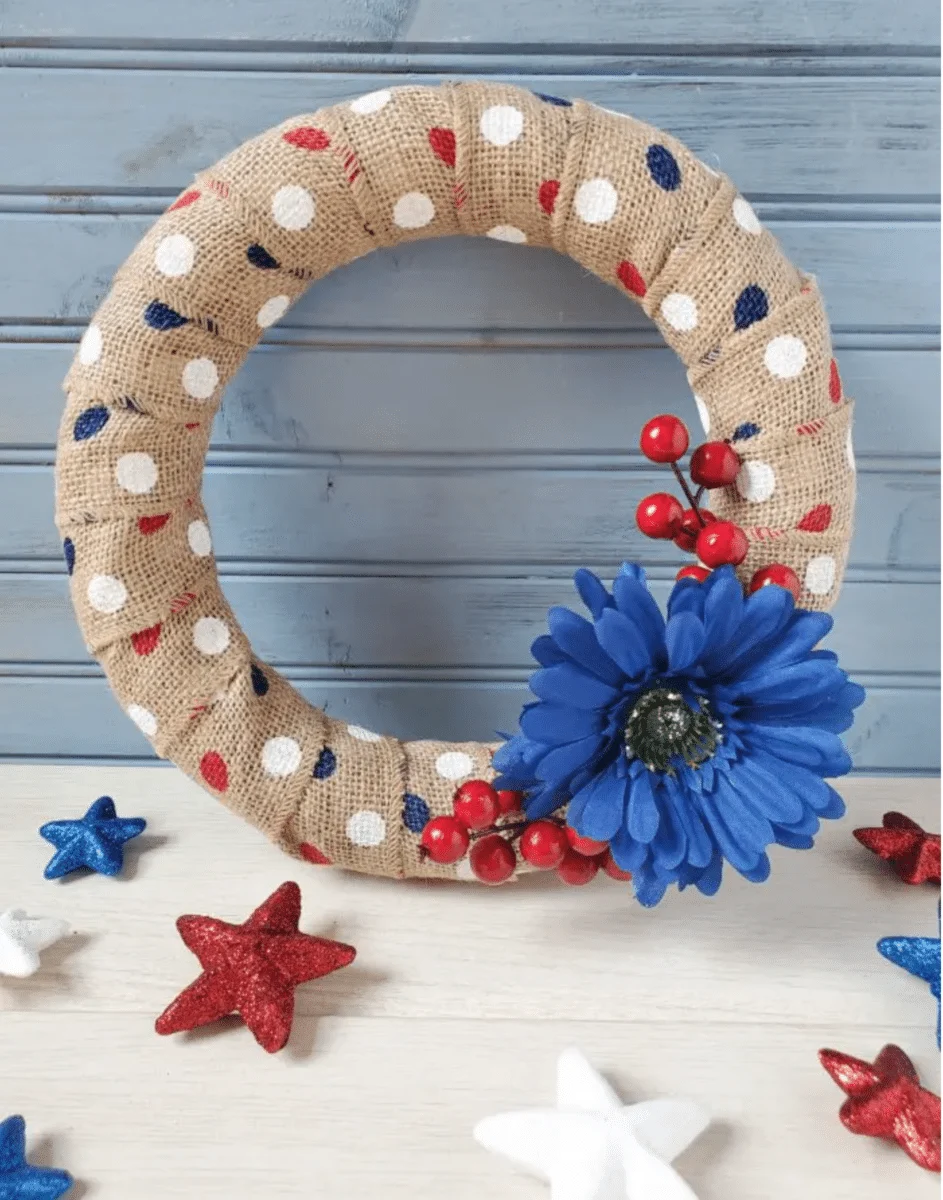 A Fourth of July burlap wreath with polka dots adorned with a blue flower and red berries, surrounded by red, white, and blue star decorations.