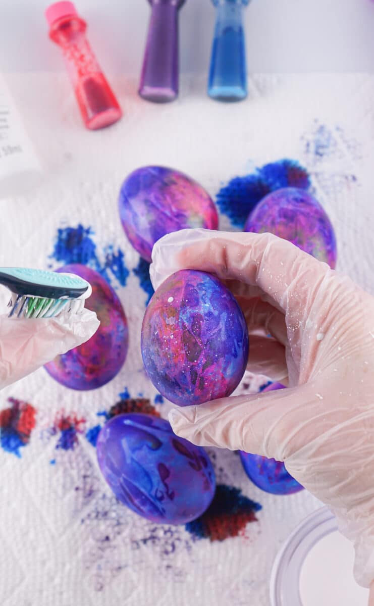 Person in gloves applying colorful dyes to a galaxy easter egg amidst a crafting session for galaxy-themed easter eggs.