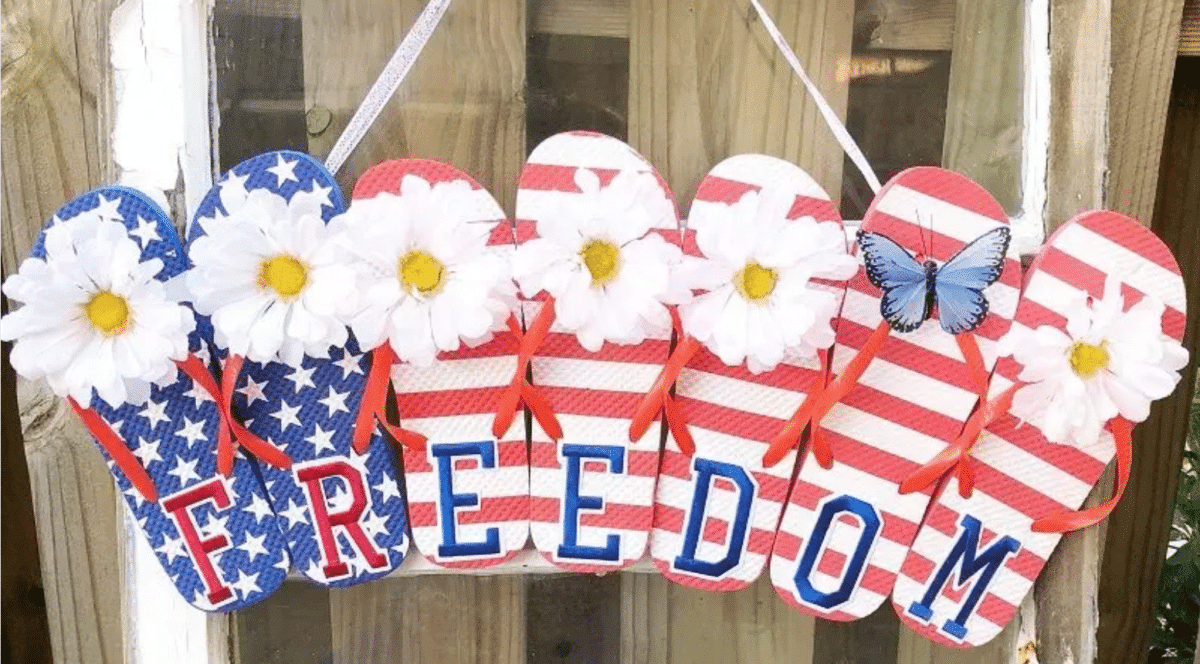 Decorative flip-flop-shaped signs with an American flag theme spelling out "freedom" adorned with white flowers and a blue butterfly, hanging on a wooden surface as part of Fourth of July wreaths