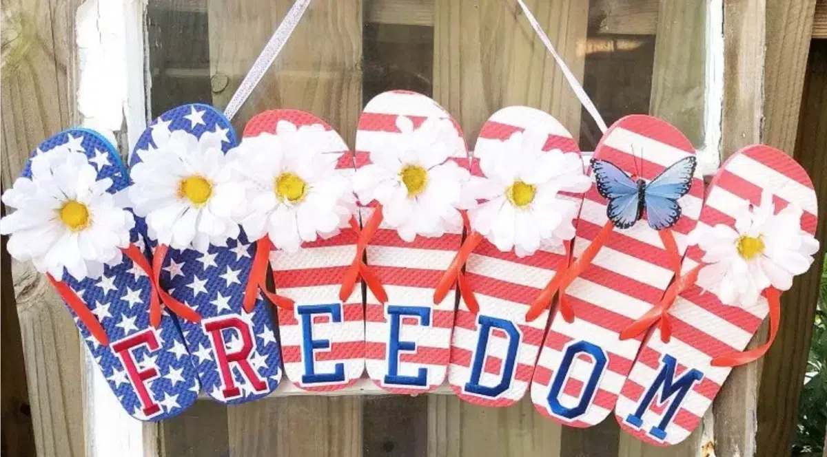 Decorative flip-flop-shaped signs with an American flag theme spelling out "freedom" adorned with white flowers and a blue butterfly, hanging on a wooden surface as part of Fourth of July wreaths