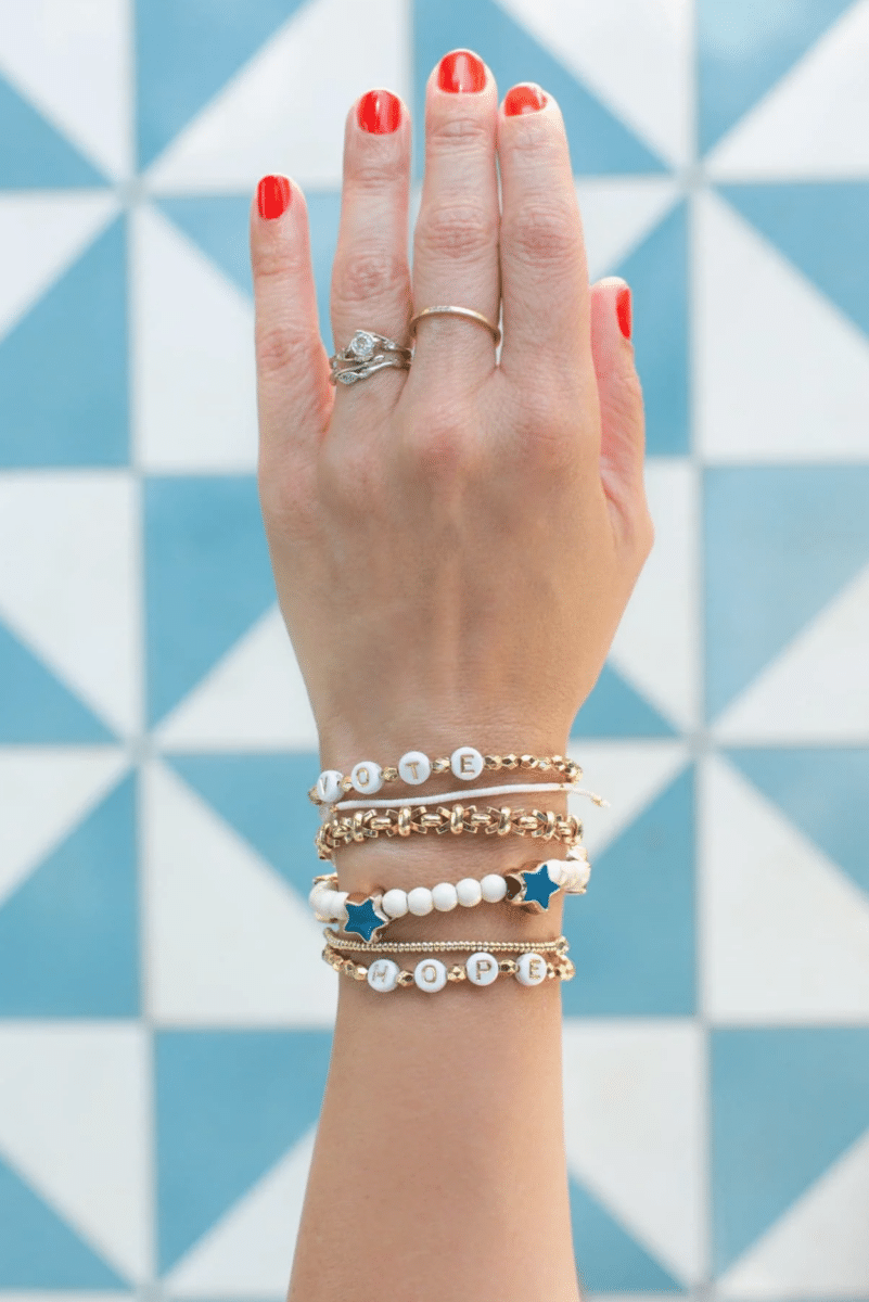 A woman's hand adorned with multiple Fourth of July crafts bracelets and a ring, set against a blue and white geometric background.