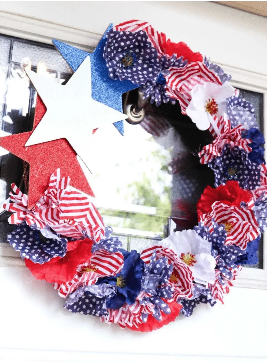 A Fourth of July-themed wreath decorated with red, white, and blue flowers and stars, displayed on a window.