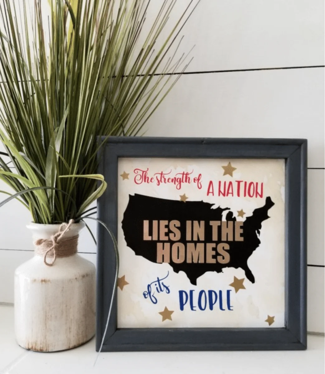 A framed quote reading "the strength of a nation lies in the homes of its people," placed next to a vase with a Fourth of July themed plant.