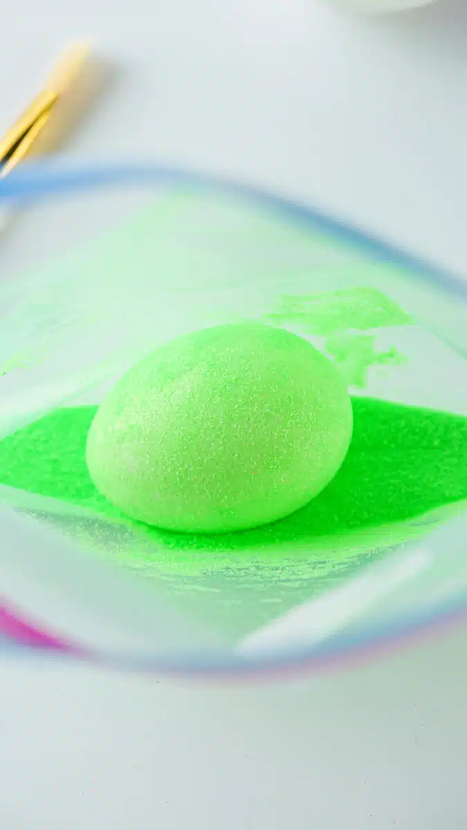 A green, glitter-covered Easter egg-shaped object on a bed of green sand, with a paintbrush in the background.